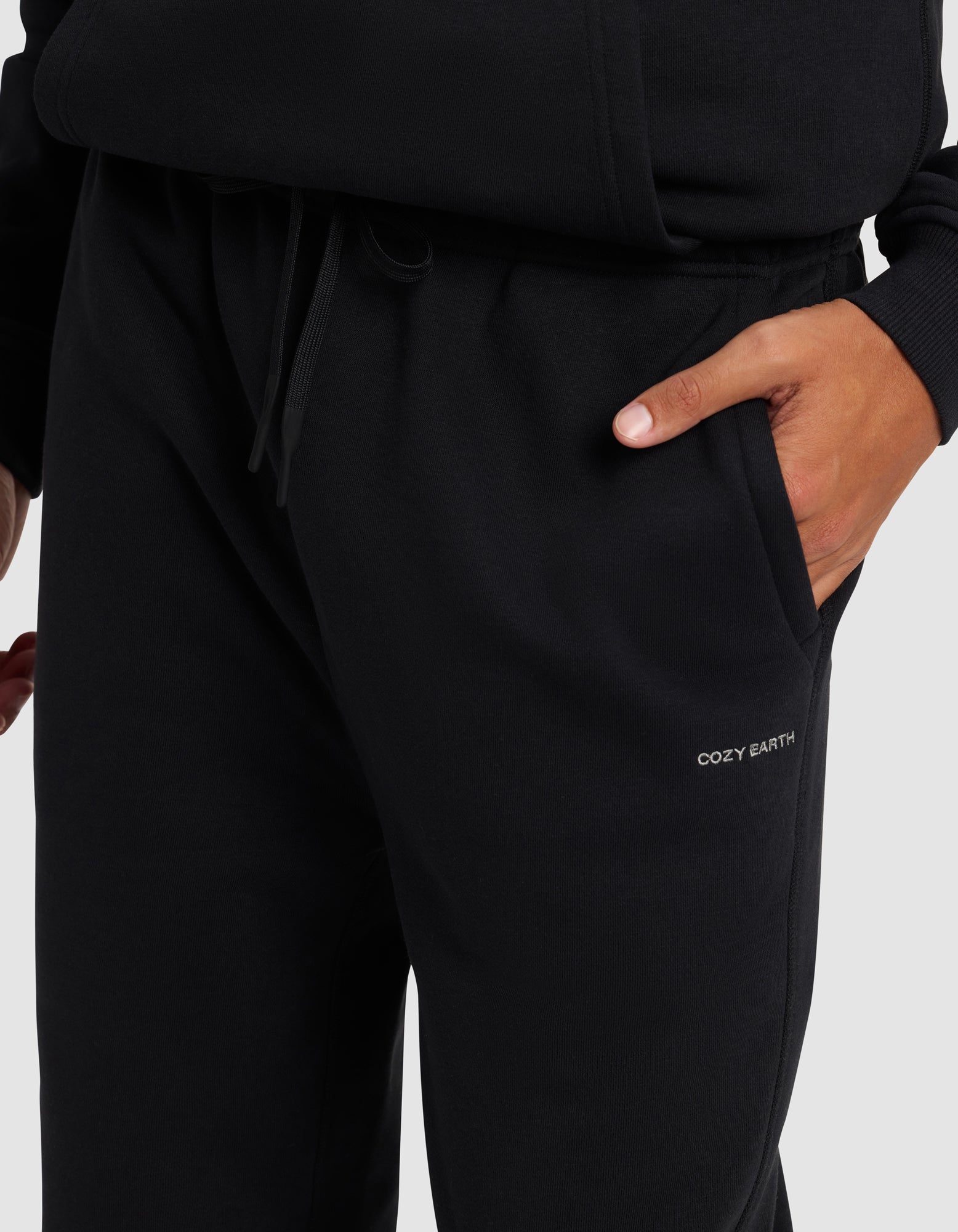 A man wears Cozy Earth Men's Cityscape 2.0 Sweatpants in black, with one hand in the pocket and "COZY EARTH" printed near it, standing against a plain light background.