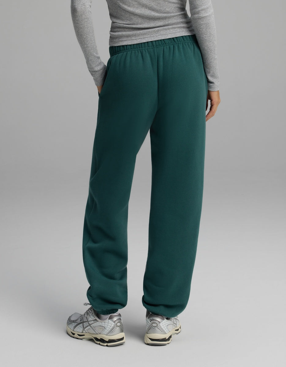 A person seen from behind wearing dark green, loose-fitting Cozy Earth Women’s CityScape Sweatpants with an elastic waistband and cuffs, paired with grey sneakers.