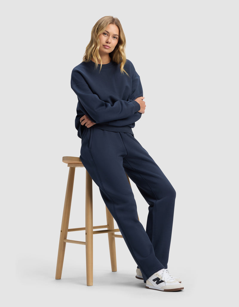 A woman with blonde hair wears the Cozy Earth Women's Cityscape 2.0 Wide Leg Pant in navy blue with a matching sweatshirt, white sneakers with gray accents, and sits on a wooden stool against a plain light gray background, looking at the camera.