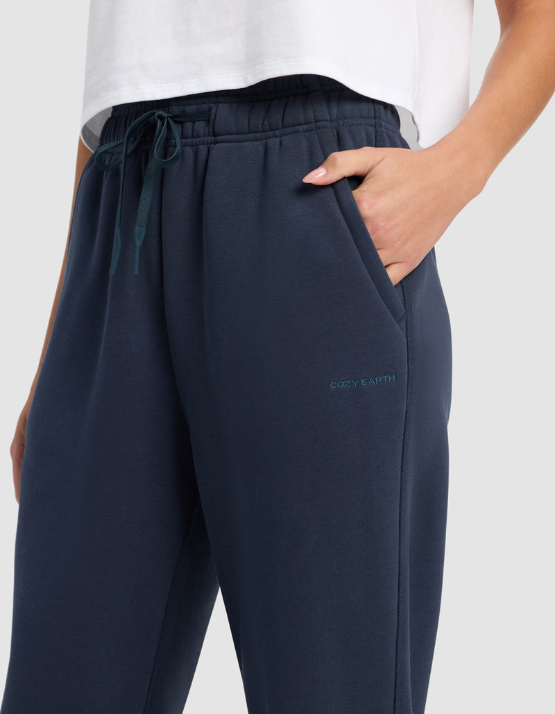 A person wears Cozy Earth Women's Cityscape 2.0 Wide Leg Pant in navy blue with a drawstring waist and "Cozy Earth" embroidered on the left thigh, paired with a white t-shirt and one hand in their pocket.