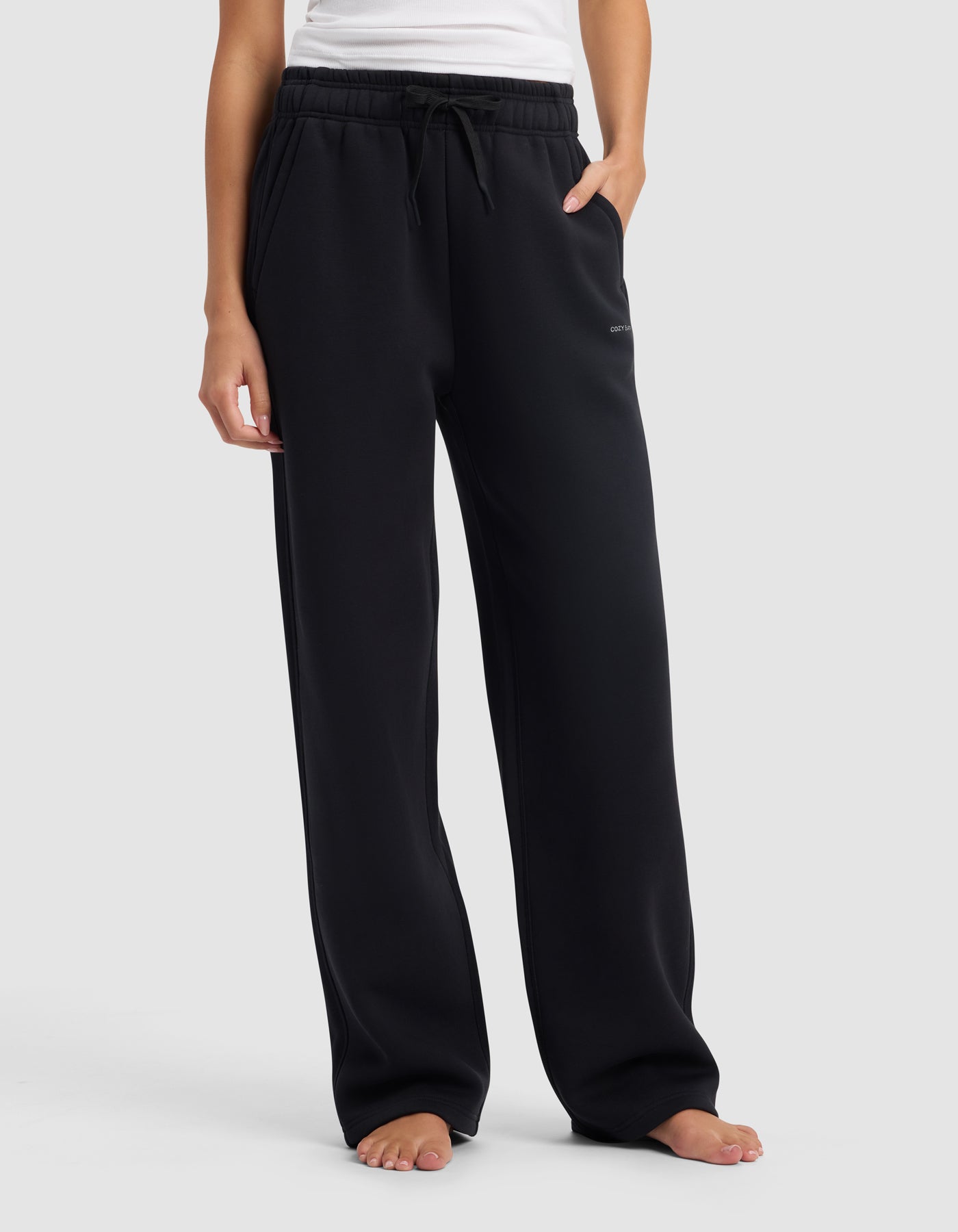 A person stands barefoot in Cozy Earth's Women's Cityscape 2.0 Wide Leg Pant, a loose-fitting black sweatpant, paired with a white sleeveless top, one hand in their pocket, against a plain light gray background.
