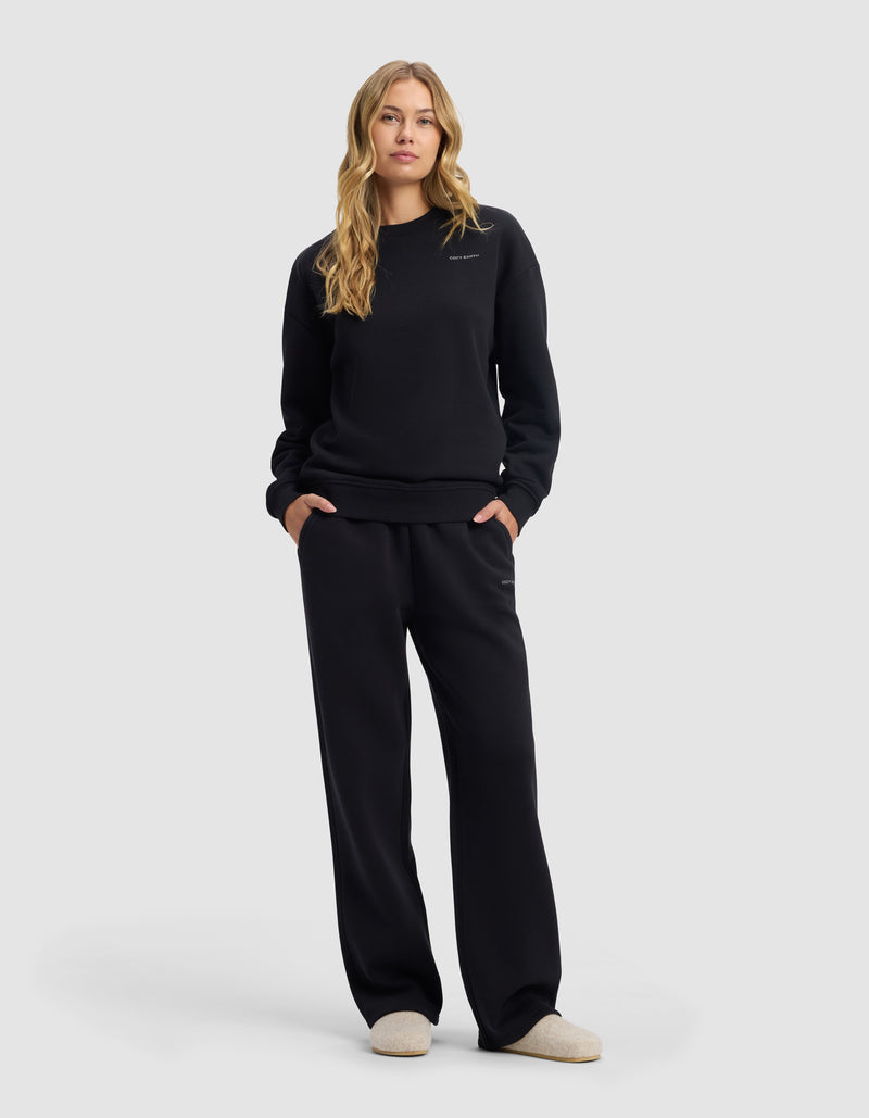 A woman with long blonde hair stands facing forward, hands in pockets, wearing Cozy Earth's Women's Cityscape 2.0 Wide Leg Pant in black, a black sweatshirt, and cream slippers against a plain light gray background.
