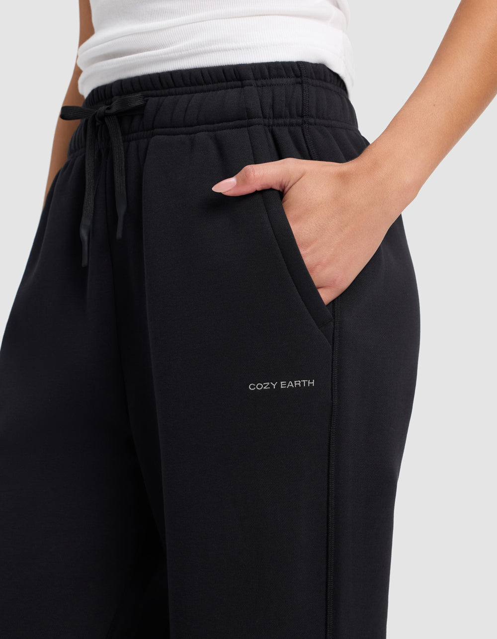 A person wears Cozy Earth Women's Cityscape 2.0 Wide Leg Pant in black, featuring "COZY EARTH" printed near the pocket, with one hand in the pocket and a white top.