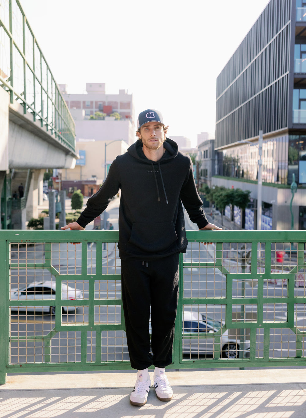 Person wearing a black hoodie and pants standing on a bridge with a cityscape background