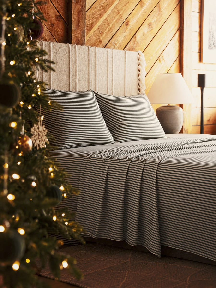 Striped green and white bedding set on a bed with a tall upholstered headboard in a rustic wood-paneled bedroom, with a decorated Christmas tree in the foreground and a lamp on a side table. 