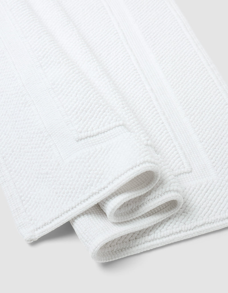 Two neatly folded Cozy Earth Classic Bath Mats, featuring a white textured surface and rectangular border design, are placed on a light gray background.