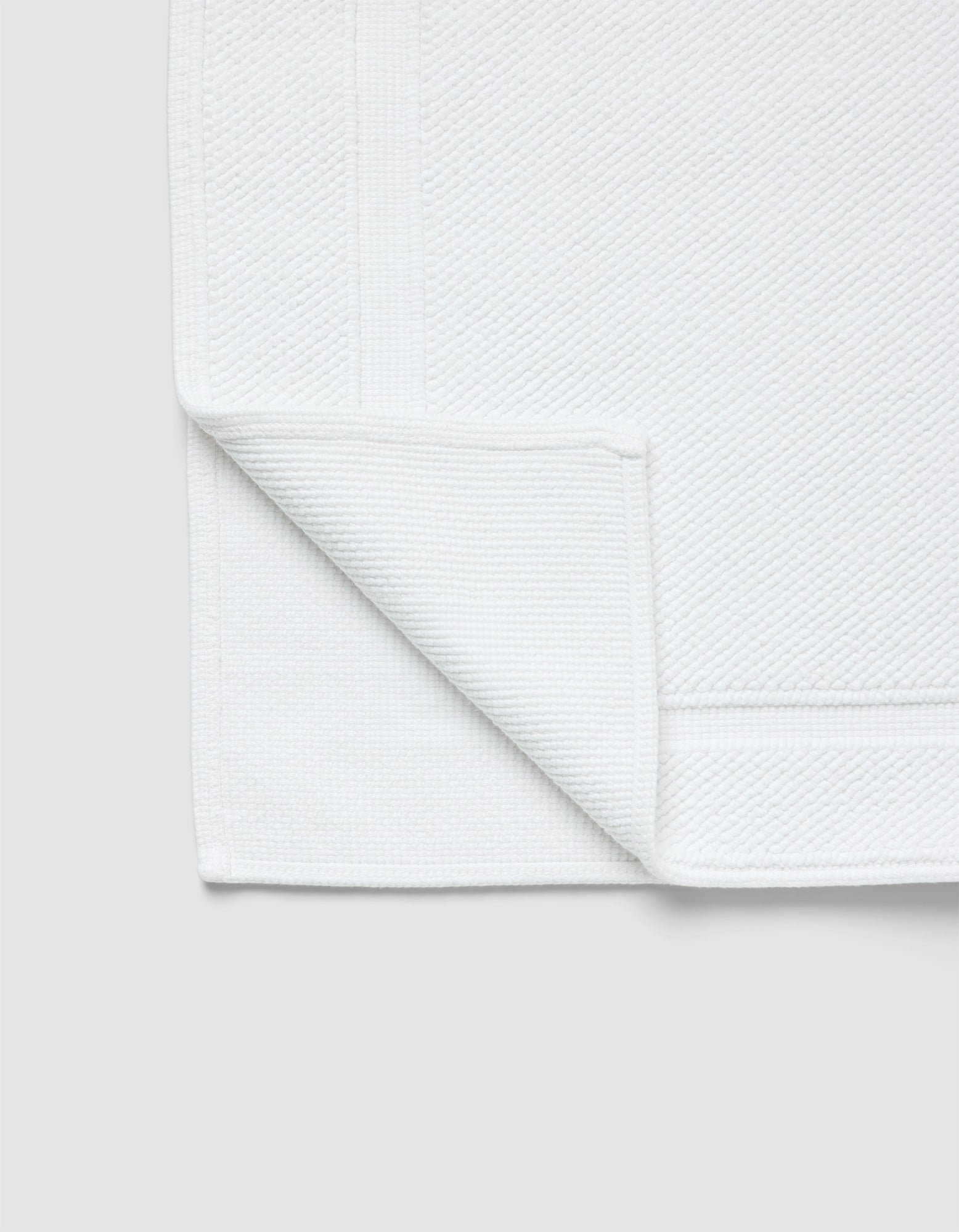 The Cozy Earth Classic Bath Mat, in white with a textured surface and simple border, is shown with one corner folded to reveal both sides against a light gray background.