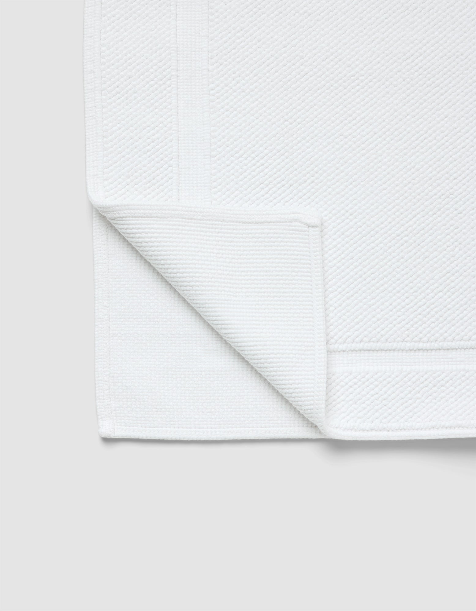 The Cozy Earth Classic Bath Mat, in white with a textured surface and simple border, is shown with one corner folded to reveal both sides against a light gray background.