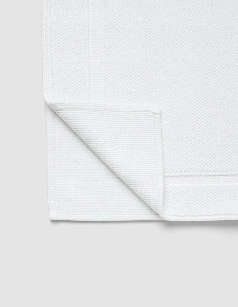 The Cozy Earth Classic Bath Mat, in white with a textured surface and simple border, is shown with one corner folded to reveal both sides against a light gray background.