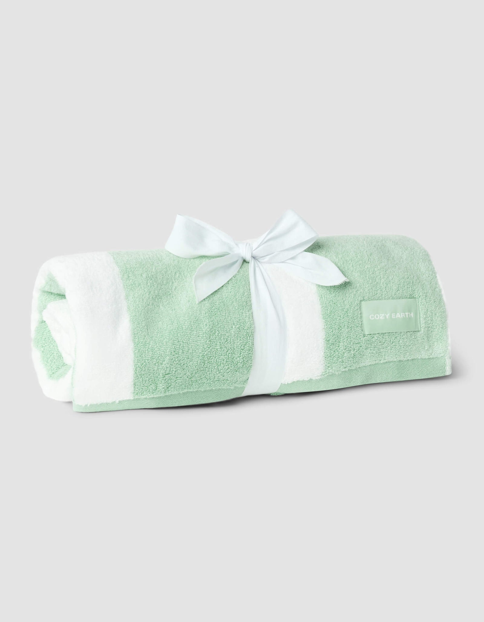 A rolled-up Classic Stripe Resort Towel by Cozy Earth in mint green and white stripes, tied with a light blue ribbon and featuring the brand’s label, is displayed against a plain light gray background.