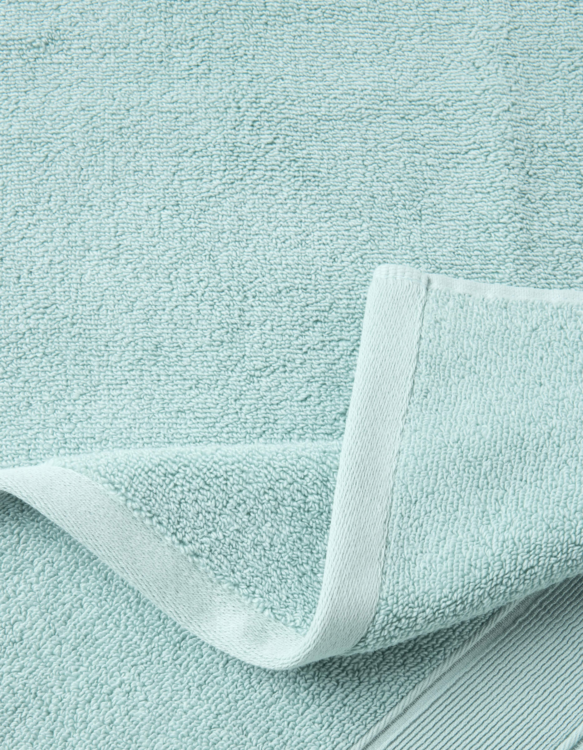 A close-up of Cozy Earth's Complete Luxe Bath Bundle in light blue shows the textured surface and neatly stitched edge, with one corner folded to display both front and back sides of the towel.|Color:Tide