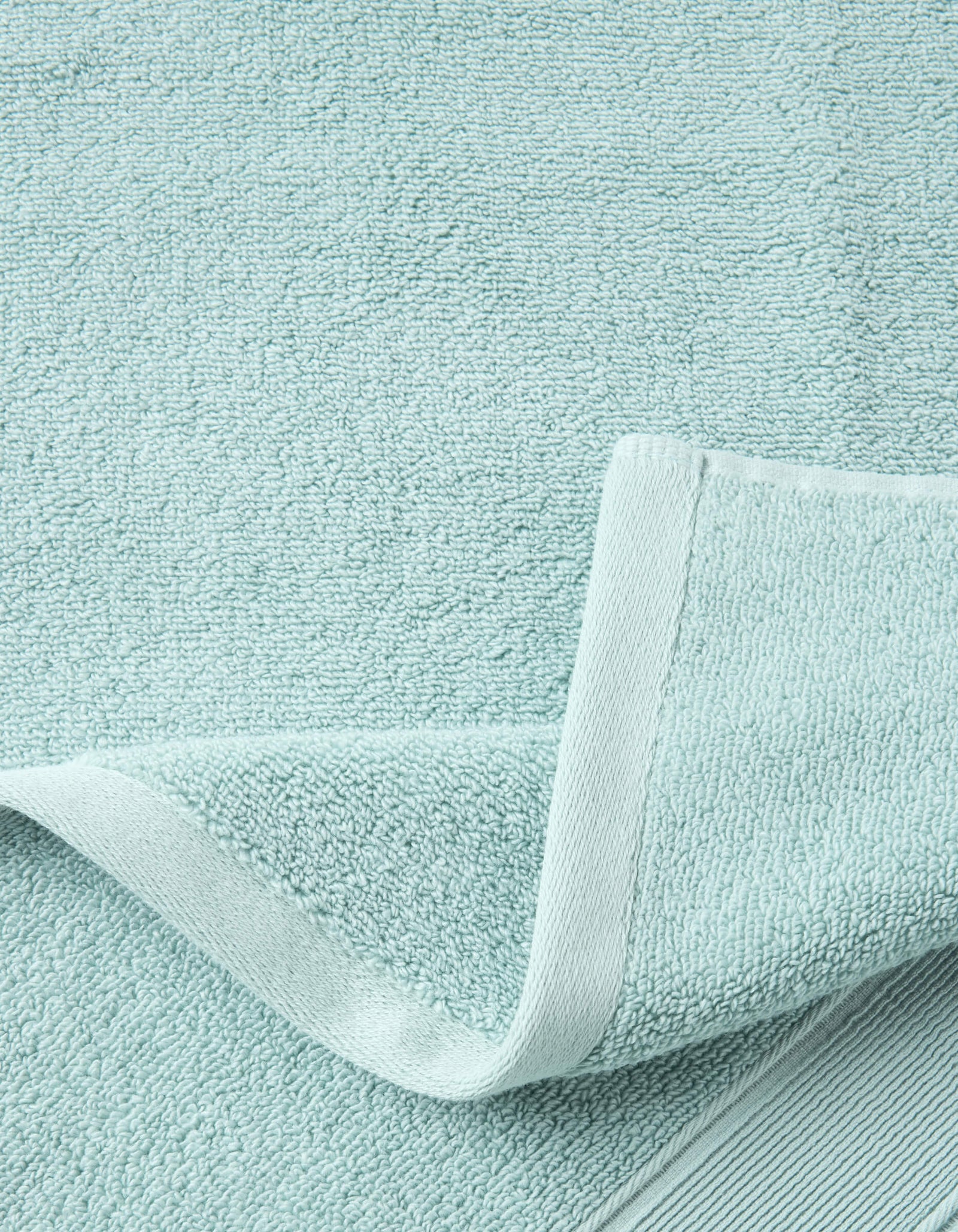 A close-up of Cozy Earth's Complete Luxe Bath Bundle in light blue shows the textured surface and neatly stitched edge, with one corner folded to display both front and back sides of the towel.