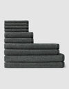 The Nantucket Bath Towel Set by Cozy Earth features a neatly stacked arrangement of dark gray towels in varying sizes, forming a stepped pattern against a plain light background.|Color:Graphite