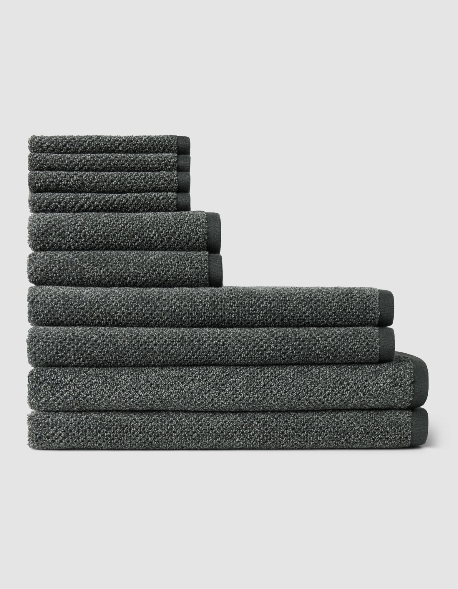 The Nantucket Bath Towel Set by Cozy Earth features a neatly stacked arrangement of dark gray towels in varying sizes, forming a stepped pattern against a plain light background.|Color:Graphite