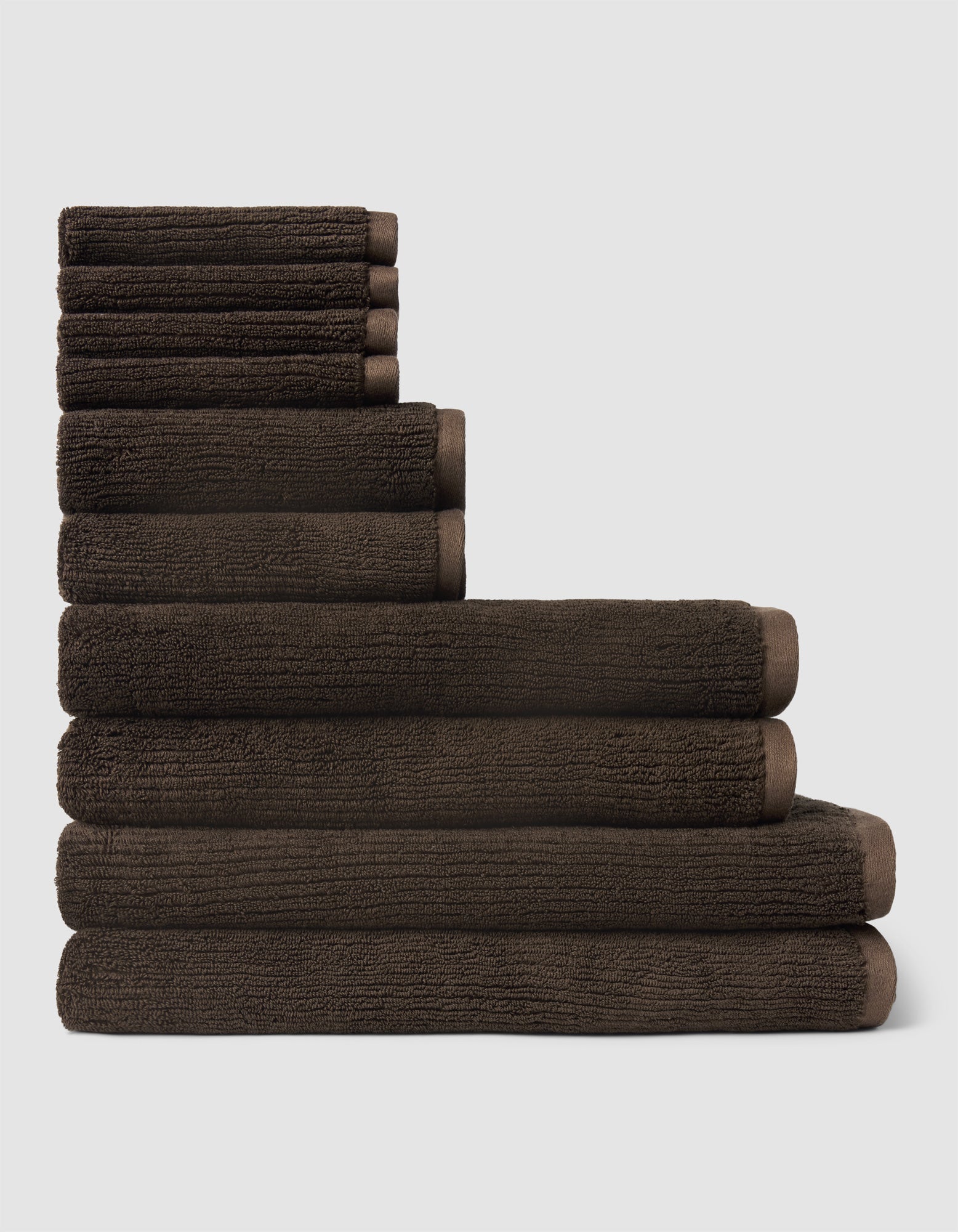 The Cozy Earth Complete Ribbed Terry Bath Bundle, featuring dark brown towels in assorted sizes, is neatly stacked from largest to smallest against a plain light gray background.|Color:Espresso