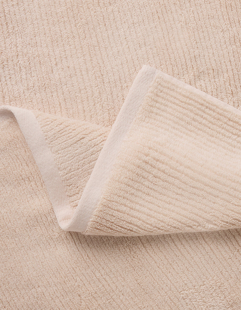 A close-up of Cozy Earth’s Complete Ribbed Terry Bath Bundle shows a folded beige towel with a soft, textured surface and subtle horizontal lines resting on a matching background.