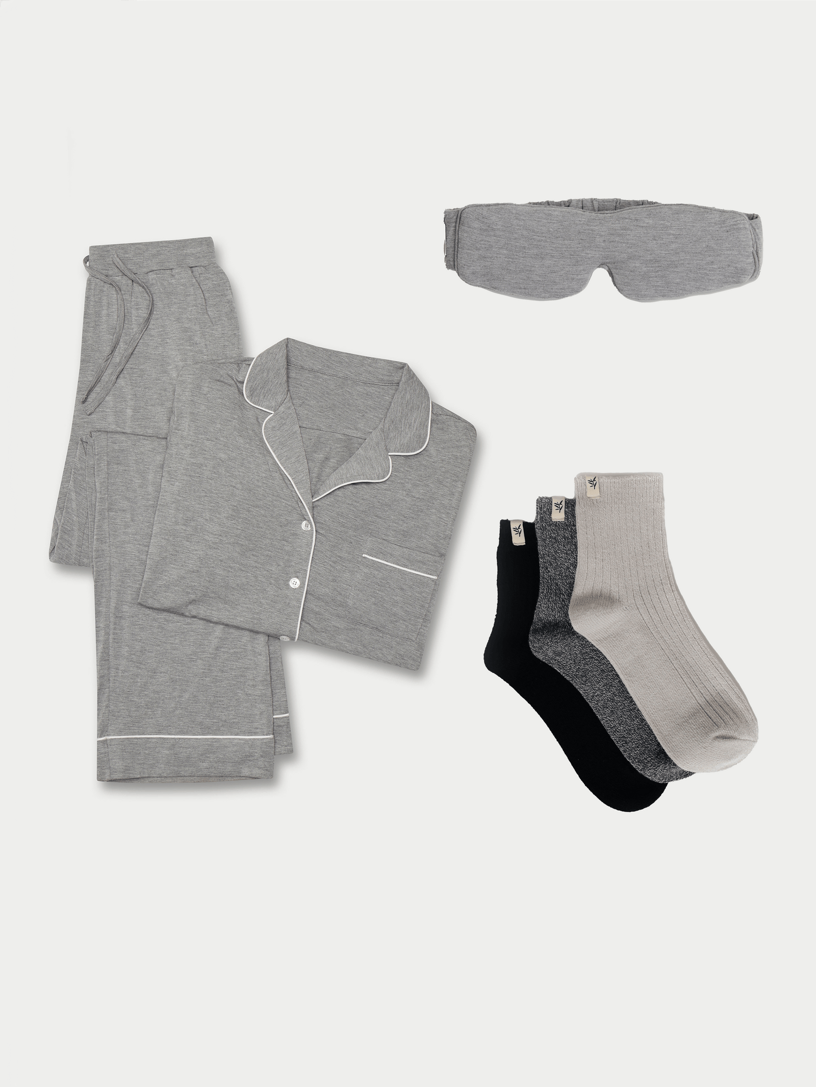 A neatly folded Women's Bedtime Bundle by Cozy Earth, which includes a set of grey pajamas featuring a button-up shirt and pants, complemented by a grey sleep mask and three pairs of socks in shades of grey, black, and light beige. All items are showcased on a plain white background. 