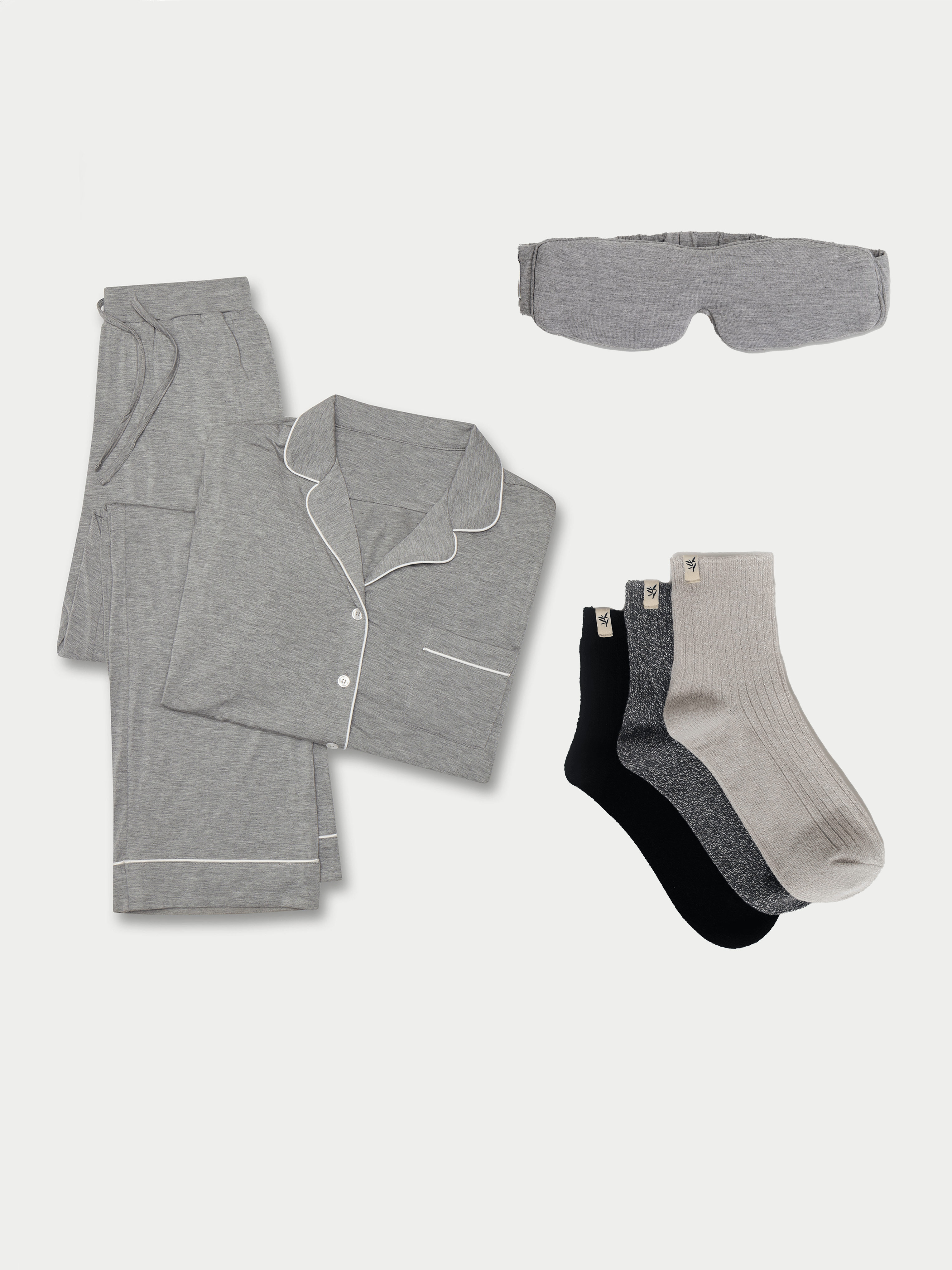 A neatly folded Women's Bedtime Bundle by Cozy Earth, which includes a set of grey pajamas featuring a button-up shirt and pants, complemented by a grey sleep mask and three pairs of socks in shades of grey, black, and light beige. All items are showcased on a plain white background. |Color:Grey