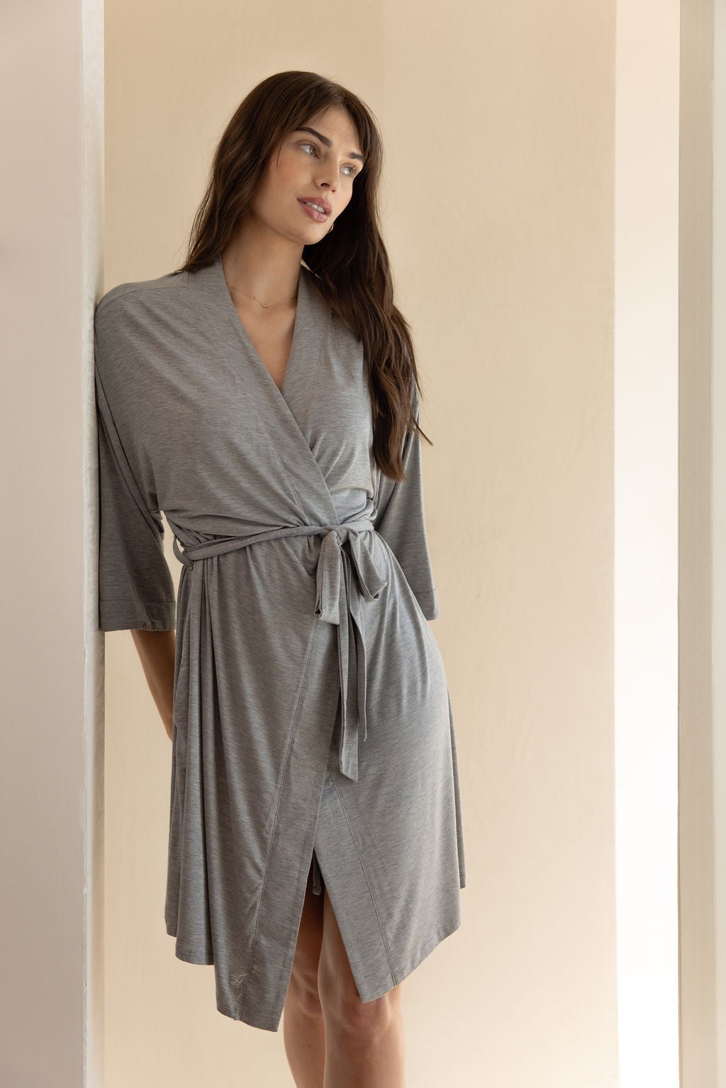 A woman with long, dark hair is wearing the Cozy Earth Women's Stretch-Knit Bamboo Kimono Robe in light gray, tied at the waist. She stands against a light-colored wall with a relaxed and pensive expression, her posture casual and her hands resting by her sides. The background is minimal and softly lit. |Color:Light Grey