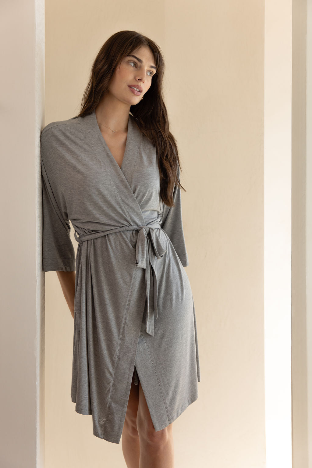 A woman with long, dark hair is wearing the Cozy Earth Women's Stretch-Knit Bamboo Kimono Robe in light gray, tied at the waist. She stands against a light-colored wall with a relaxed and pensive expression, her posture casual and her hands resting by her sides. The background is minimal and softly lit. |Color:Grey