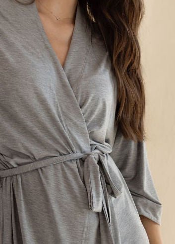 A person with long hair is wearing a Cozy Earth Women's Stretch-Knit Bamboo Kimono Robe in gray, tied at the waist with a matching belt. The robe features a V-neckline and three-quarter length sleeves. The background is neutral beige. The person's face is not visible in the image. 