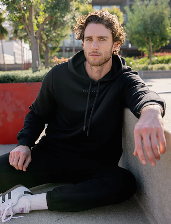 Man with curly hair sitting outdoors on a concrete surface, wearing a black hoodie and black pants. His white sneakers have black stripes. Green foliage and a red wall are visible in the background, suggesting an urban park setting.