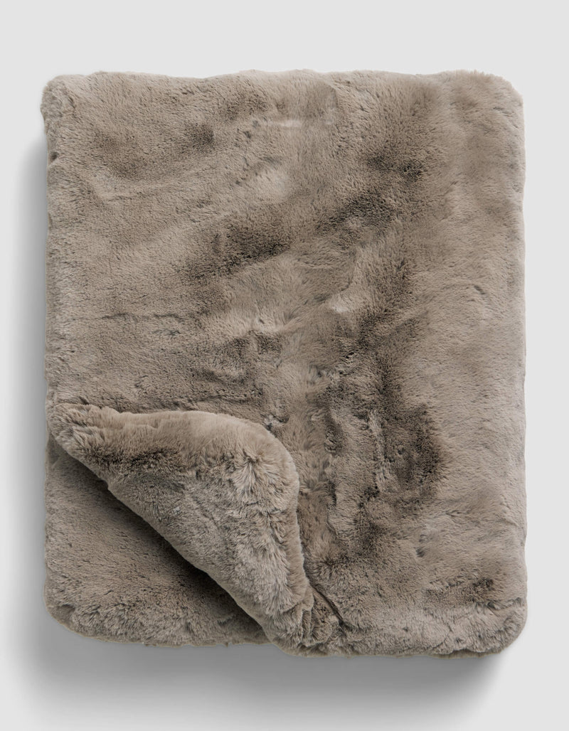The Superplush Cuddle Blanket by Cozy Earth is a soft, light brown faux fur blanket with a plush texture, shown folded on a plain light gray background. A corner is turned up to reveal its fluffy underside.