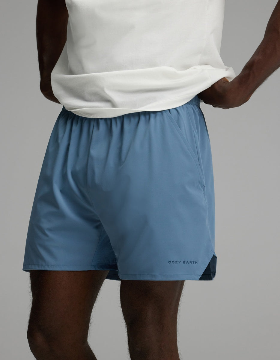 A person wearing a white shirt and Cozy Earth Men’s Day & Night Short 5″ in Nordic Blue, which features the brand’s name on the left leg, standing with hands on hips against a plain background.