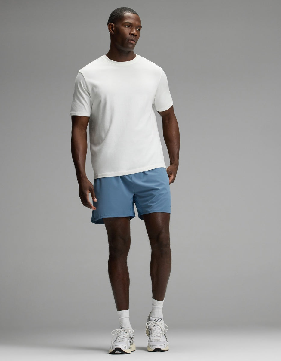 A man stands against a plain background wearing a white T-shirt, Cozy Earth Men’s Day & Night Short 5″ in blue, white socks, and white sneakers.