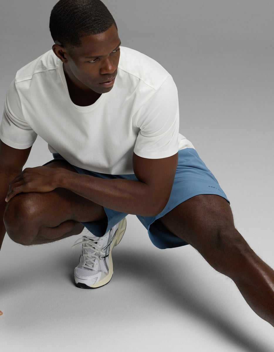 A man wearing Cozy Earth’s Men’s Day & Night Short 5″ in blue, paired with a white t-shirt and white sneakers, is crouching on one knee and looking to the side against a light grey background.