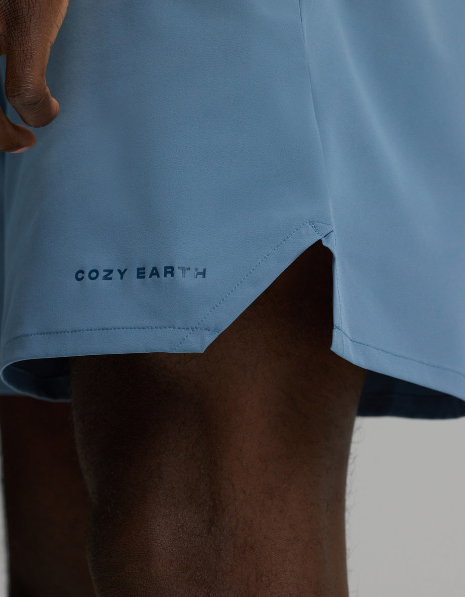 A close-up of a person wearing the Men’s Day & Night Short 5″ by Cozy Earth in light blue, featuring the brand’s name printed near a small side slit on the hem. Only the lower part of the shorts and part of the person’s leg are visible.