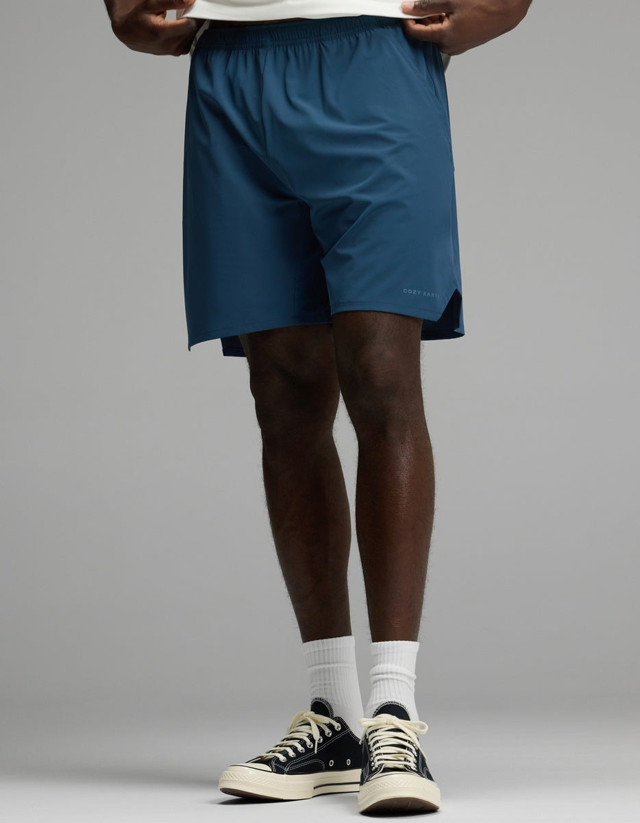 A person is shown from the waist down wearing Cozy Earth’s Men’s Day & Night Short 7″ in Fathom, paired with white socks and black sneakers, standing against a plain gray background.|Color:Fathom