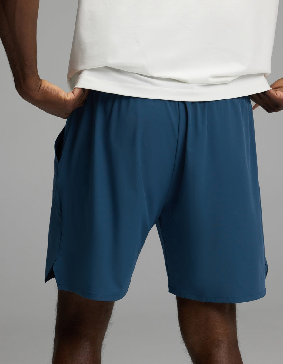 A person wearing Cozy Earth’s Men’s Day & Night Short 7″ in blue shorts and a white shirt stands with hands on hips against a plain background.
