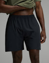 A person wearing a green shirt and the Cozy Earth Men’s Day & Night Short 7″ in Jet Black stands with one hand on their hip against a plain gray background.|Color:Jet Black
