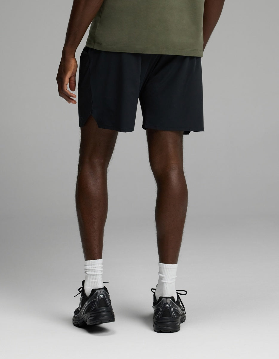 A person stands facing away, wearing Cozy Earth’s Men’s Day & Night Short 7″ in black, paired with a green shirt, white socks, and black sneakers against a plain, light background.