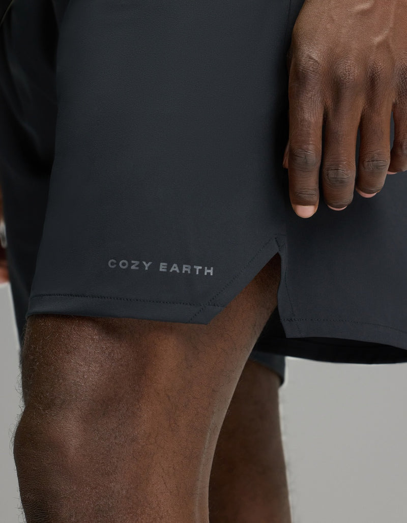 A close-up of a person’s hand resting near the hem of the Cozy Earth Men’s Day & Night Short 7″, featuring “COZY EARTH” printed above a side slit.