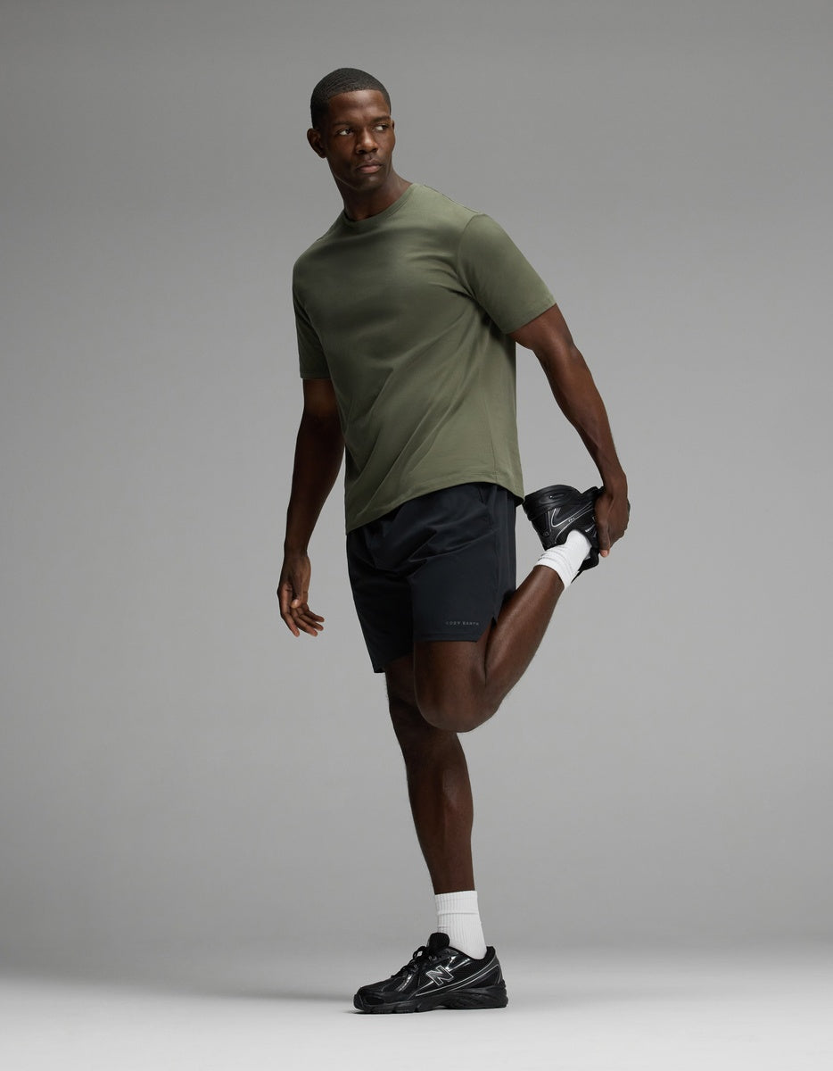 A man wearing Cozy Earth’s Men’s Day & Night Short 7″ in black and an olive green t-shirt stands on one leg, holding his ankle behind him in a quad stretch. He pairs the shorts with white socks and black sneakers, gazing off to the side against a gray background.