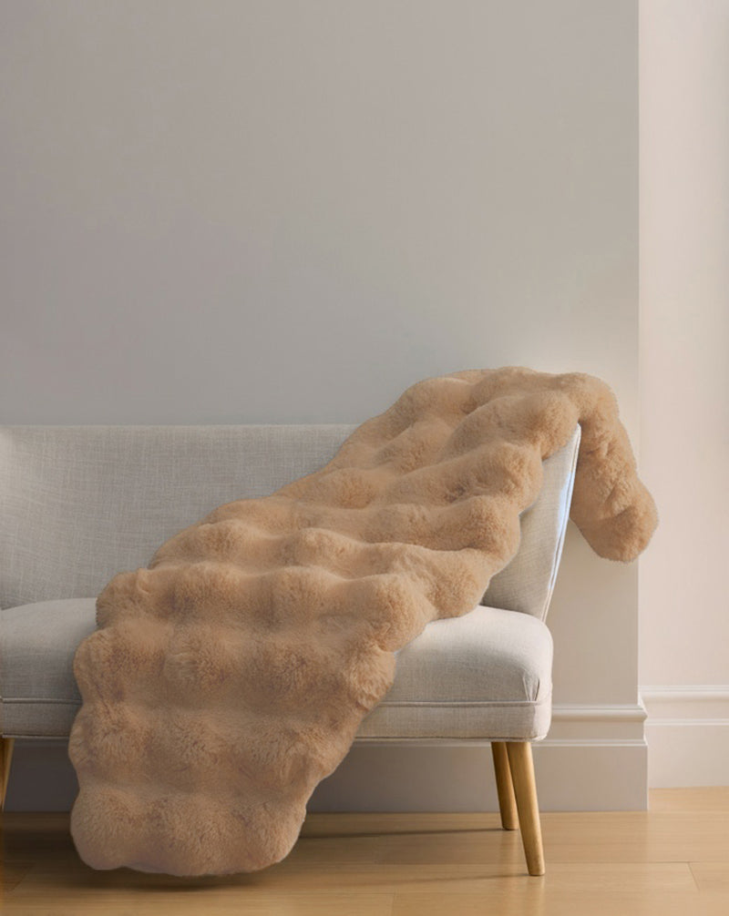 The Cozy Earth Double Bubble Cuddle Blanket, a light beige faux fur throw with a wavy texture, is draped over the arm of a modern light gray sofa in a minimalist room with light wood floors and neutral walls.