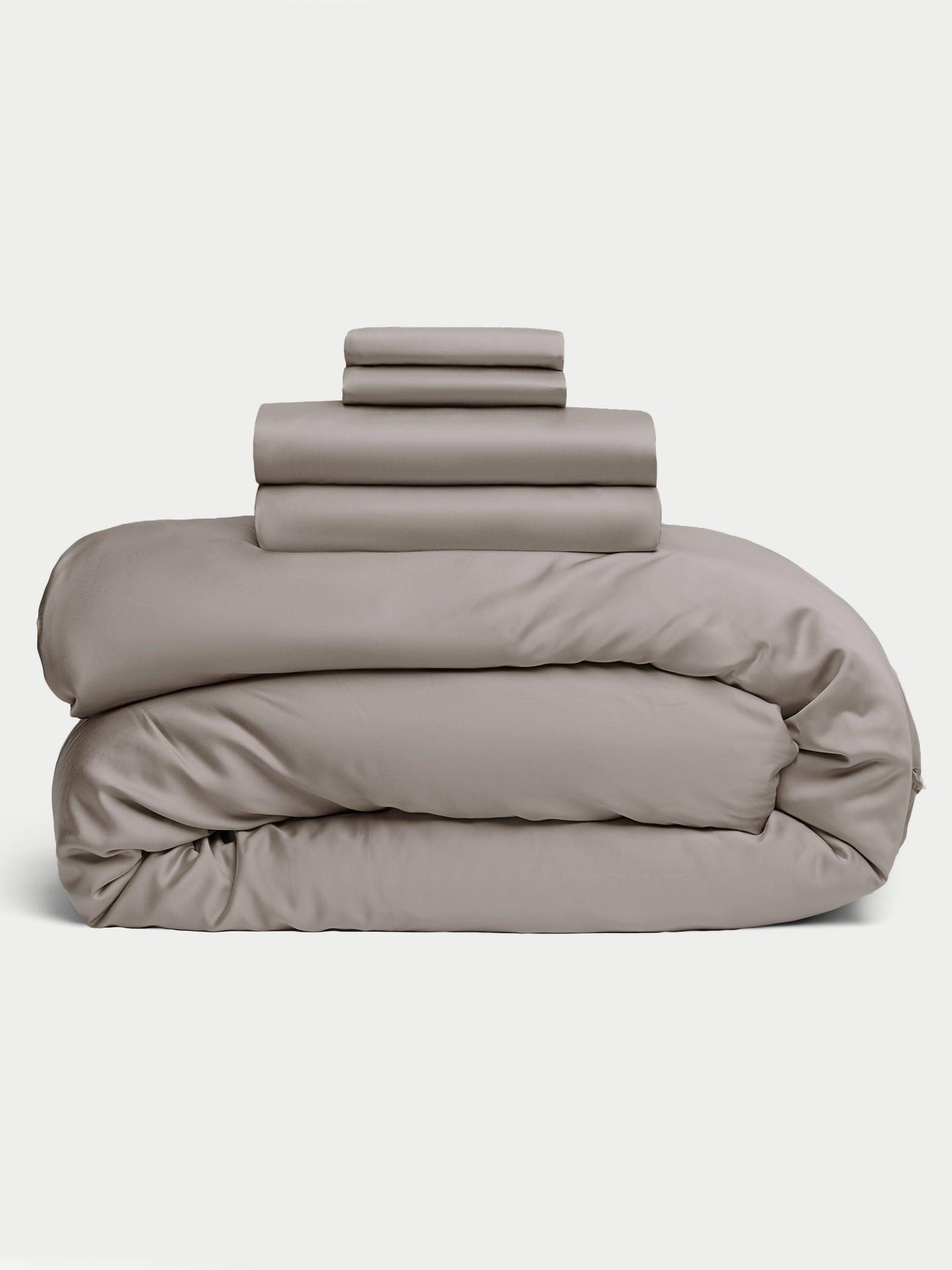 The Bamboo Bedding Deluxe Bundle by Cozy Earth is beautifully arranged with a large comforter at the bottom, topped by stacked sheets and pillowcases in light gray, all presented against a white background. 