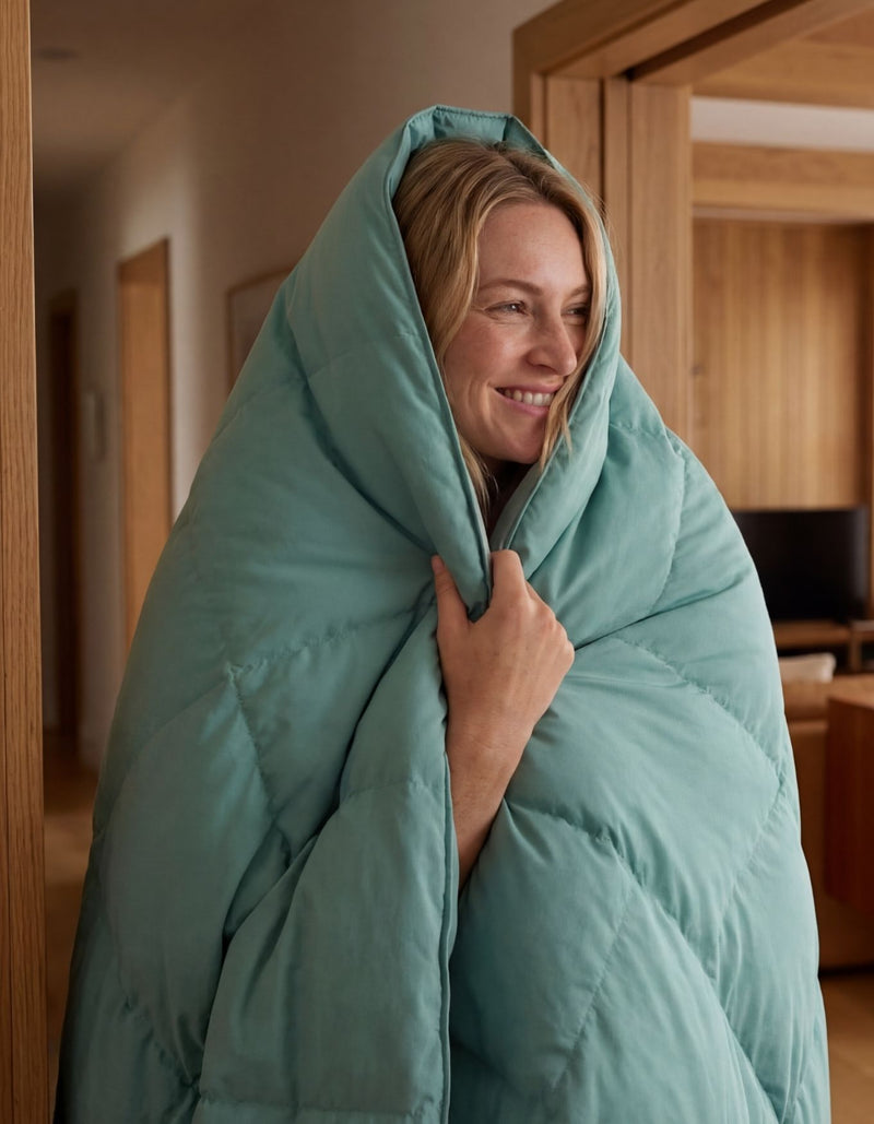 A smiling person stands indoors, wrapped in the Cozy Earth Down Alternative Throw, with wooden door frames and a softly lit room in the background.