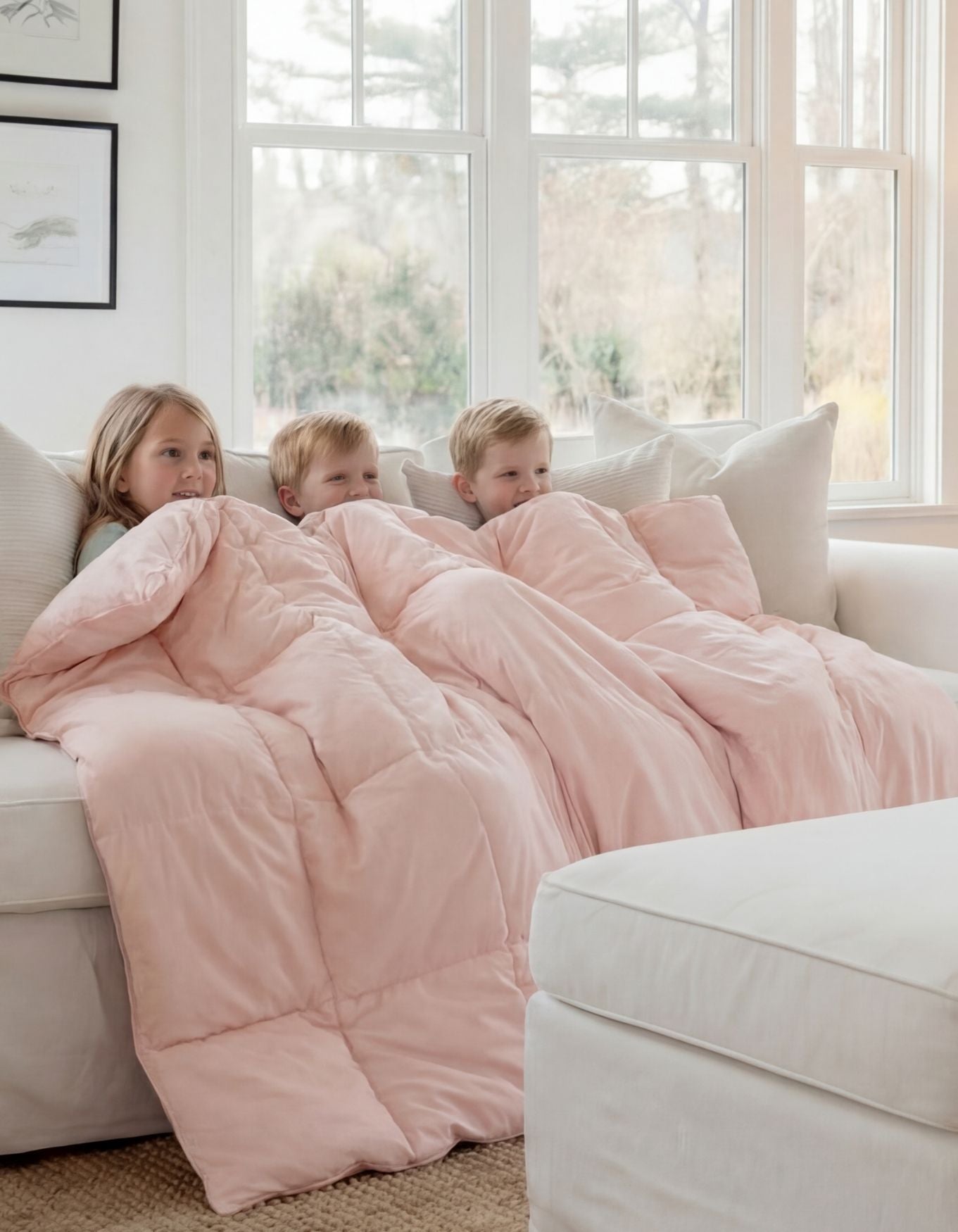 Three young children sit on a white couch, wrapped together under the Cozy Earth Down Alternative Throw in light pink. They look cozy as they relax in front of large windows bathed in natural light. 
