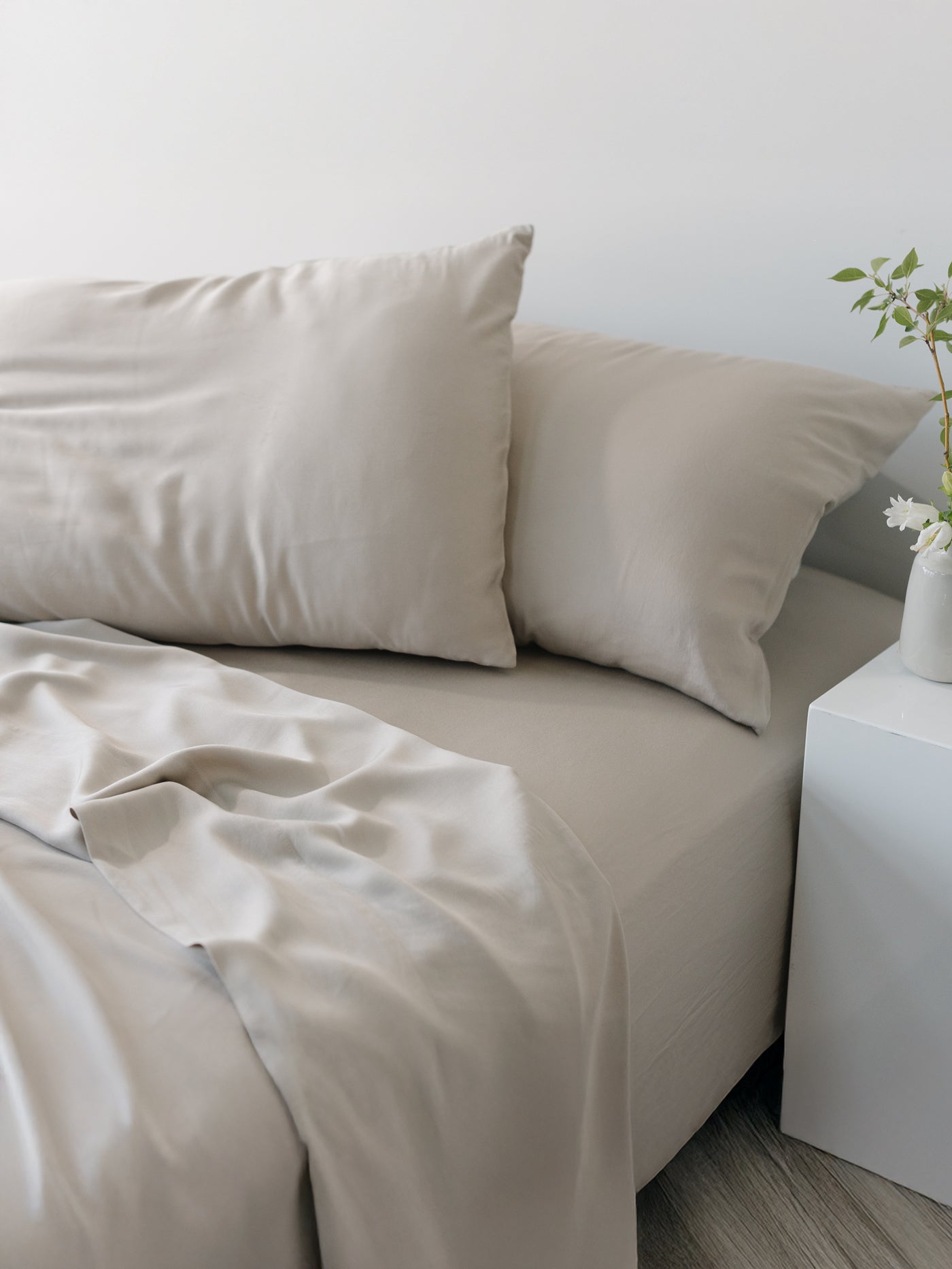 A neatly made bed featuring the Bamboo Sheet Set by Cozy Earth, in beige, is next to a white side table. A small vase with green plant stems is placed on the table, creating a calm and minimalist bedroom setting. |Color:Driftwood