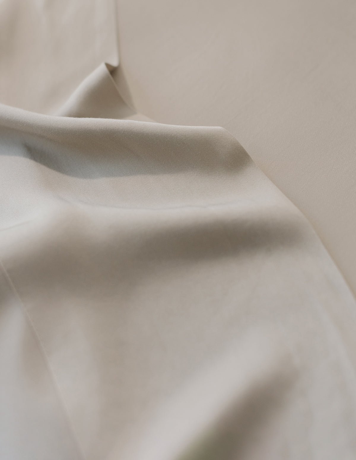 The Bamboo Sheet Set by Cozy Earth is shown in a close-up display, featuring smooth beige fabric crumpled elegantly on a flat surface. Gentle folds and shadows enhance its soft texture, silky feel, and subtle sheen. 