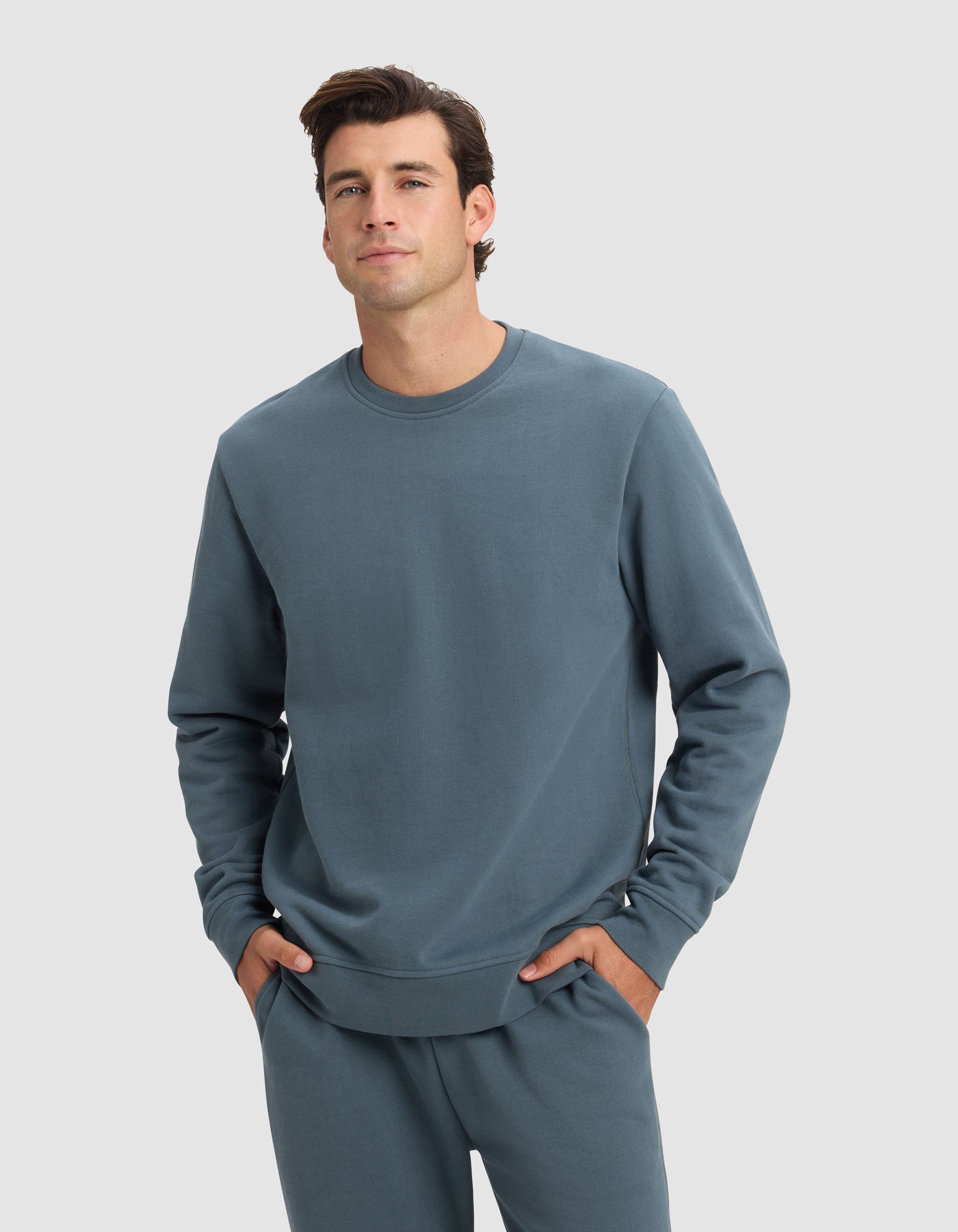 A man stands against a plain background, wearing the Cozy Earth Men's Dylan Crewneck in blue-gray with matching sweatpants, hands in his pockets and a neutral expression.