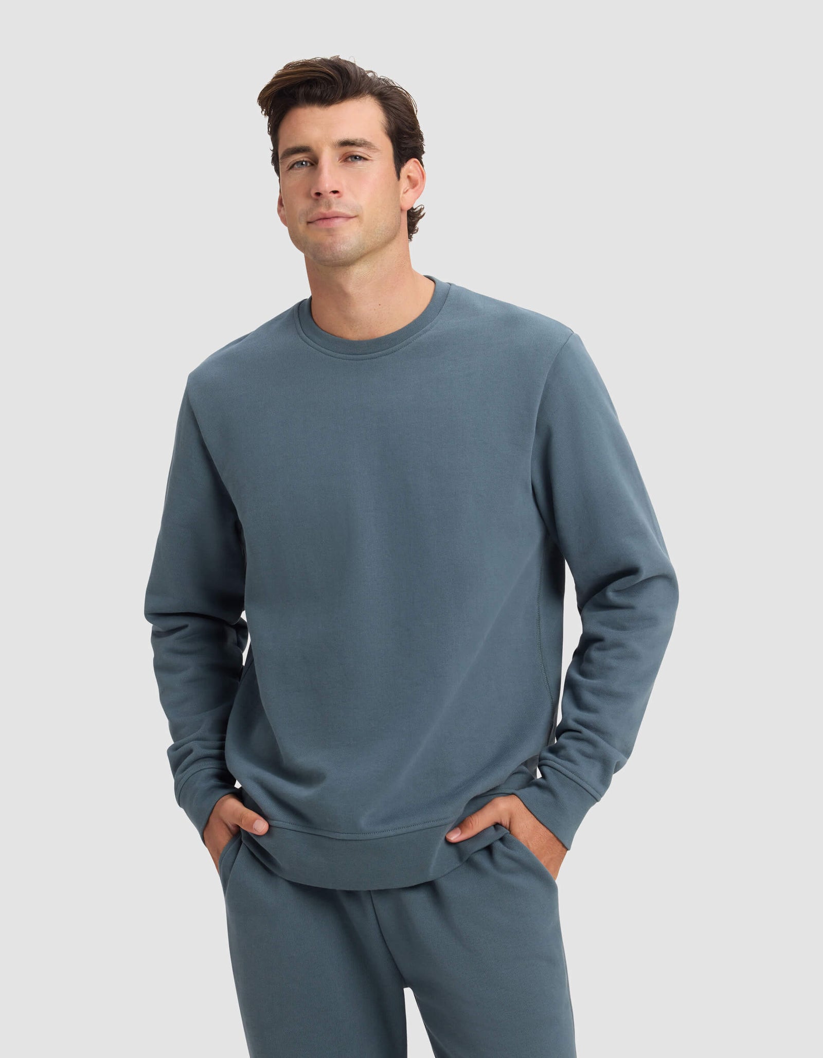 A man wears the Cozy Earth Men's Dylan Crewneck in blue-gray, standing with hands in pockets and facing the camera against a plain light gray background.