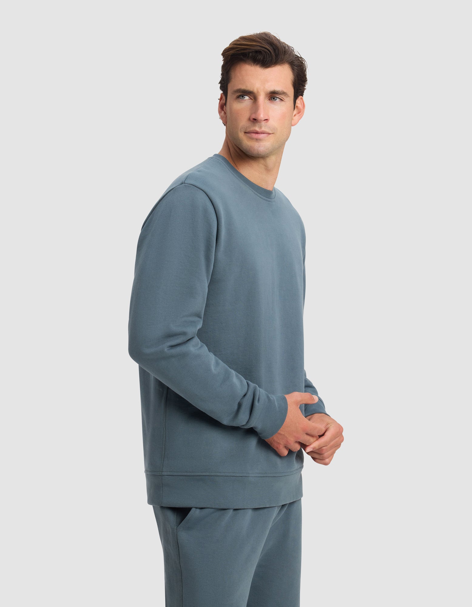 A man wearing the Cozy Earth Men's Dylan Crewneck in blue stands against a plain light gray background, looking slightly to his left with a neutral expression.|Color:Dawn