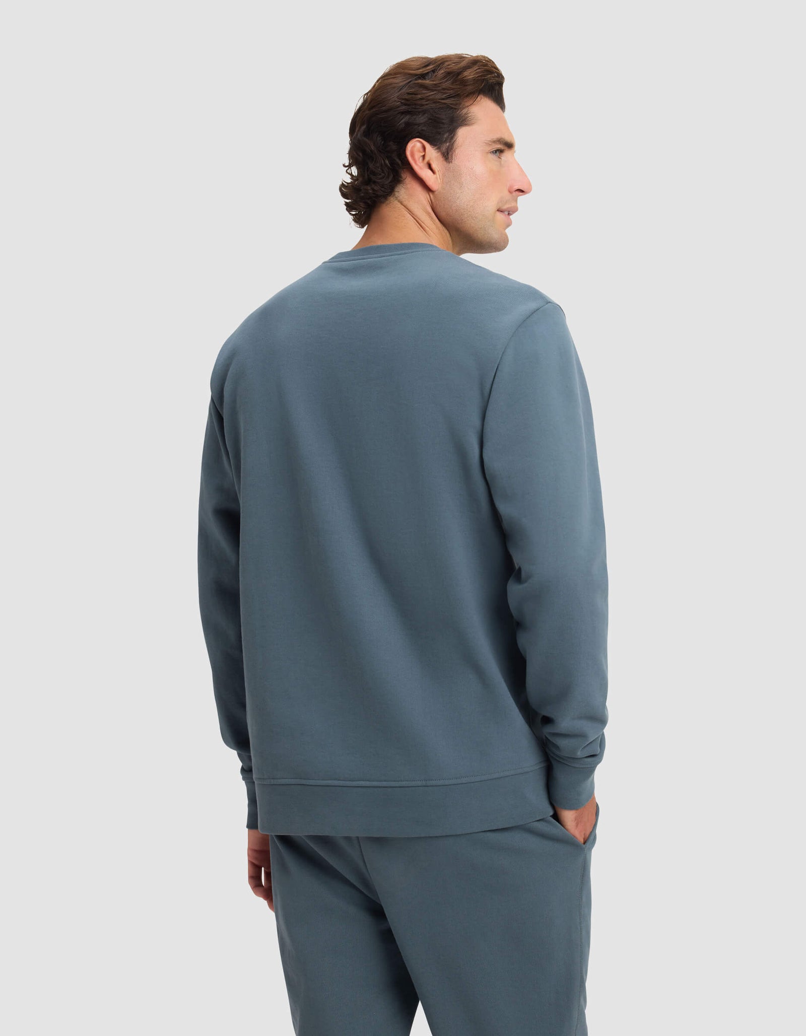 A man with short brown hair wears the Cozy Earth Men's Dylan Crewneck in blue, paired with matching sweatpants, standing against a plain light gray background and glancing to the side.