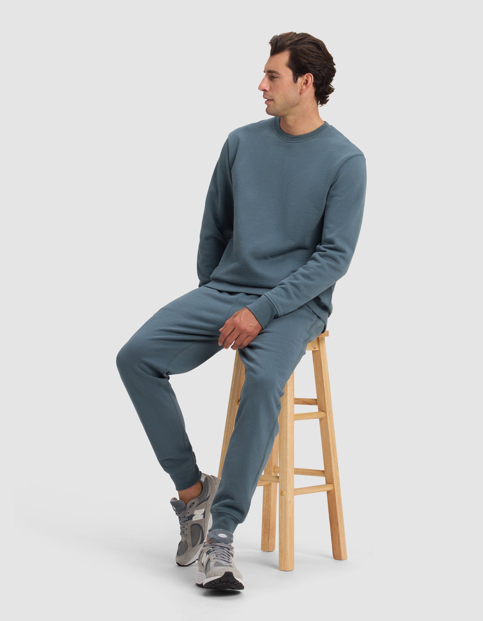 A man wearing the Cozy Earth Men's Dylan Crewneck and matching sweatpants sits on a wooden stool, looking to the side. He pairs the outfit with gray sneakers against a plain, light-colored background.|Color:Dawn