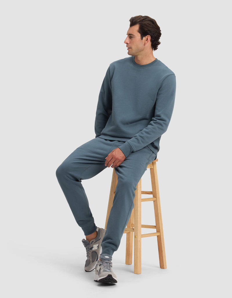 A man wearing the Cozy Earth Men's Dylan Crewneck and matching joggers sits on a wooden stool, looking to his right. He pairs the outfit with gray sneakers against a plain, light-colored background.