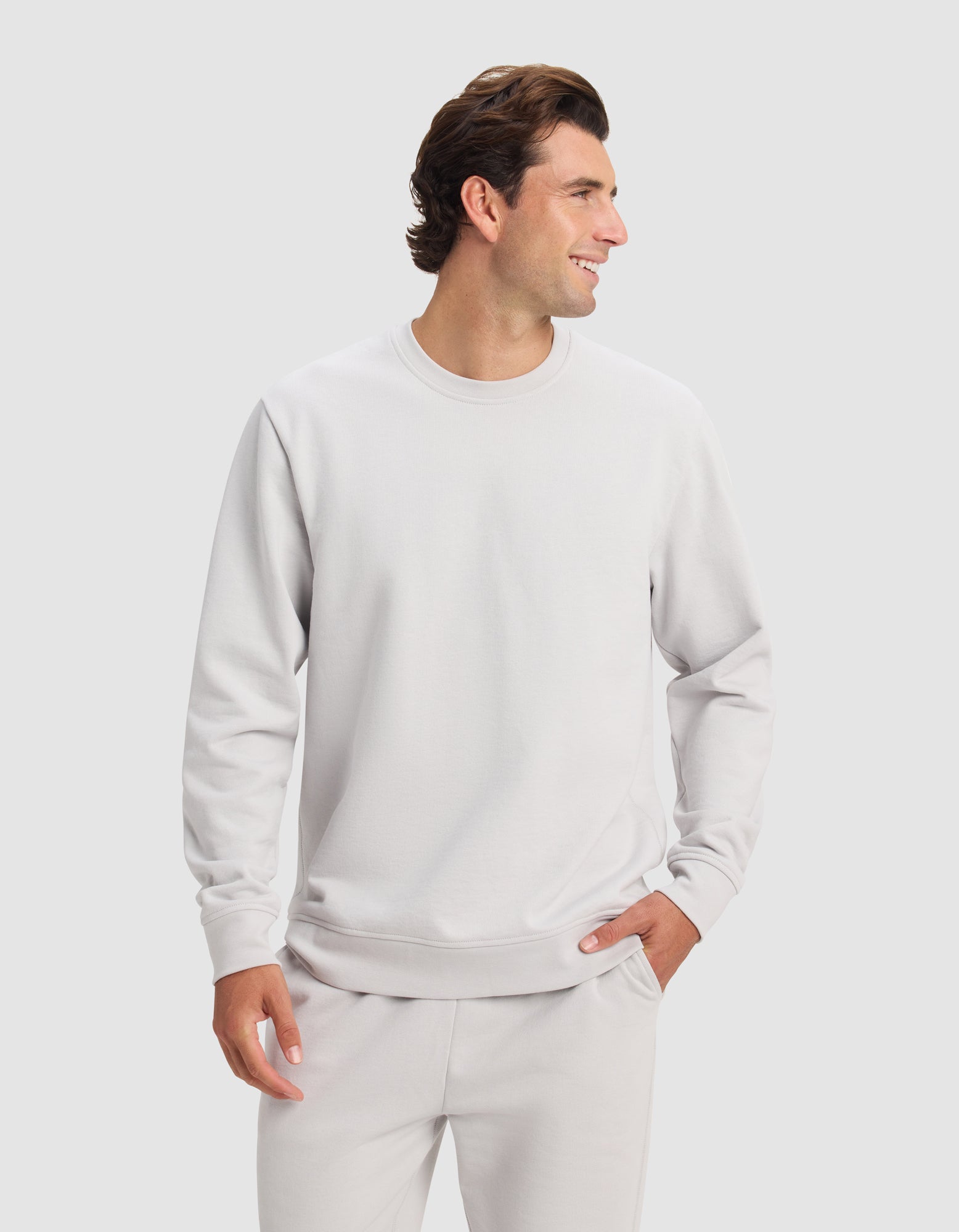 A man wears the Cozy Earth Men's Dylan Crewneck and matching pants, standing against a light background with one hand in his pocket, smiling and looking to his left.